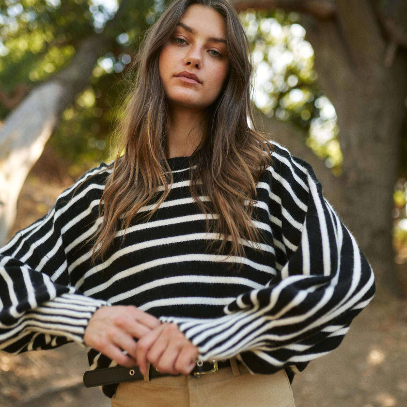 Woman wearing a black and white striped sweater and beige skirt standing outdoors with trees in the background