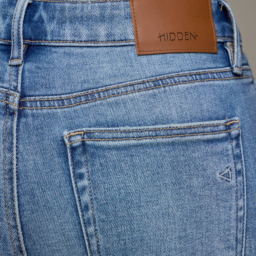 Close-up of blue jeans with a brown leather patch and 'Hidden' branding.