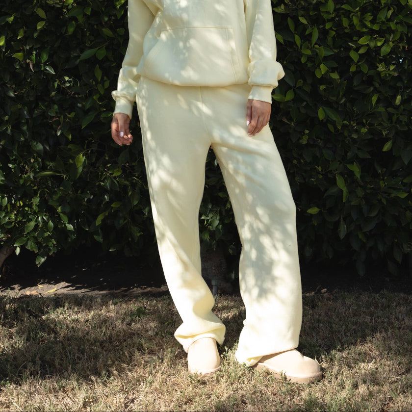 Person wearing a cream-colored hoodie and pants standing against a green hedge.