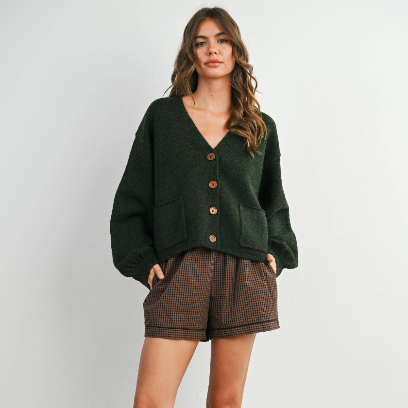 Woman wearing a green cardigan, brown shorts, and black boots against a white background