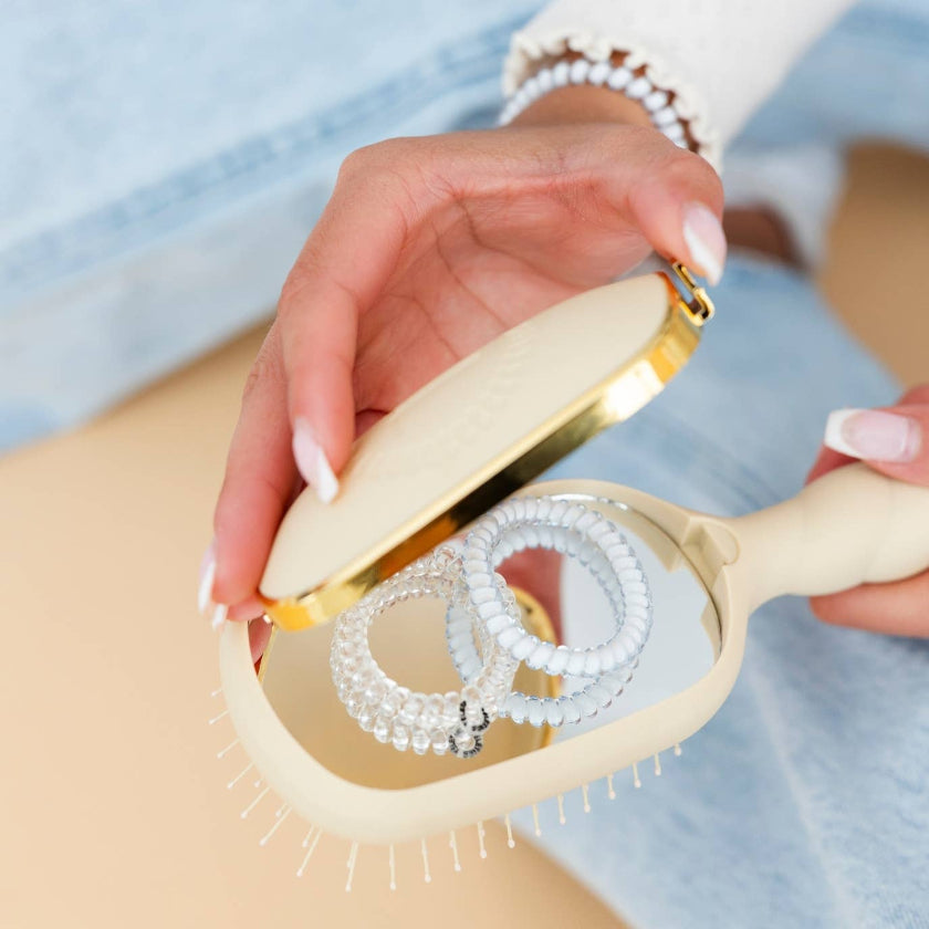 Hand holding a beige hairbrush with clear bristles against a light blue background