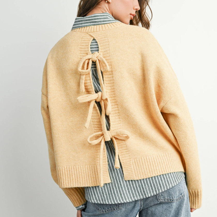 Woman wearing a mustard yellow cardigan with bow details on a plain background