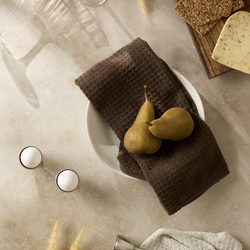 Brown waffle-textured towel on a white plate with pears, on a beige surface with glasses and bread.
