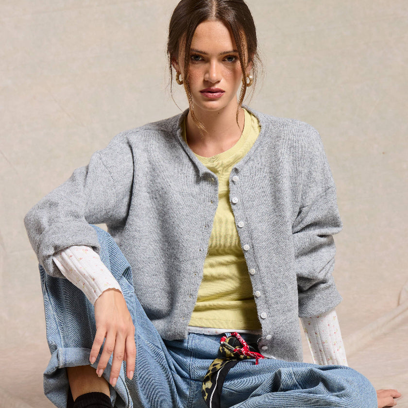 Woman sitting on a beige surface wearing a gray cardigan, yellow shirt, blue jeans, and yellow sneakers.