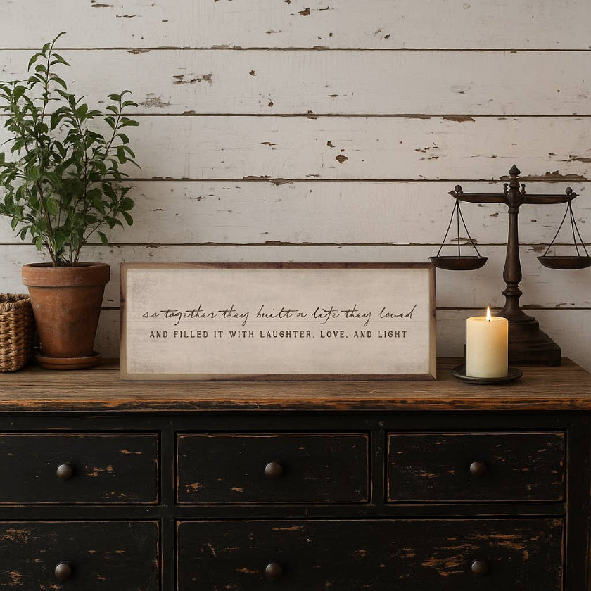Decorative setup with a framed quote, candle, and scale on a wooden dresser against a rustic wall.