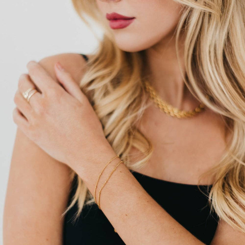 Woman wearing gold jewelry including a necklace, bracelet, and ring on a plain background