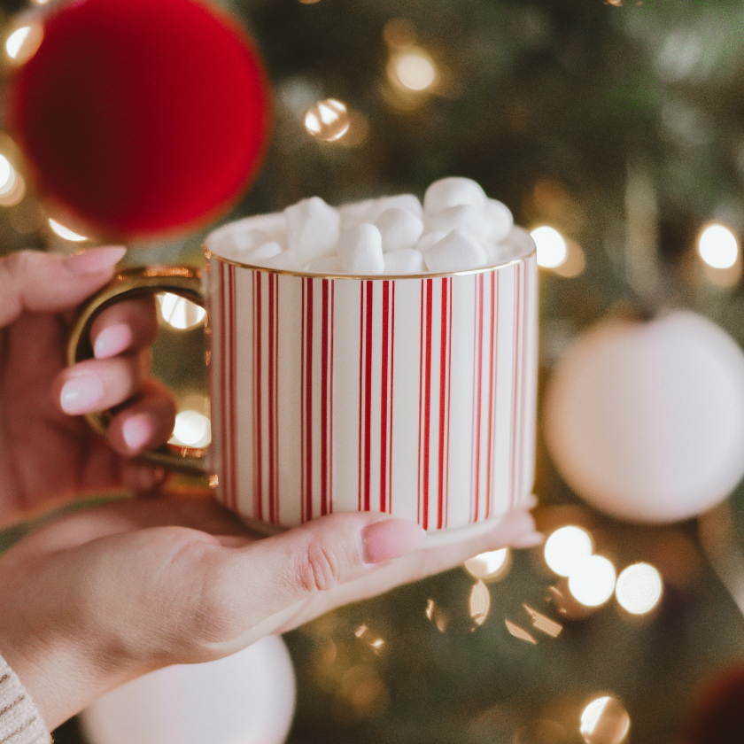 Hand holding a mug with red and white stripes filled with hot chocolate and marshmallows, against a festive Christmas tree background.