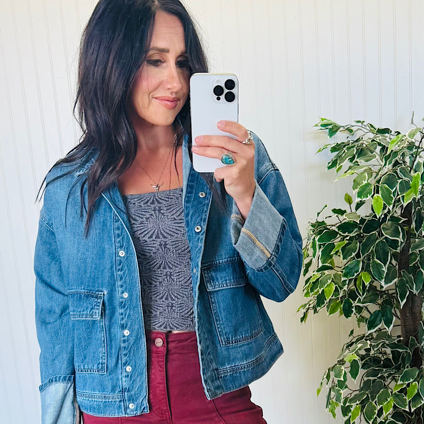 Woman taking a selfie wearing a denim jacket and patterned top, standing next to a plant.