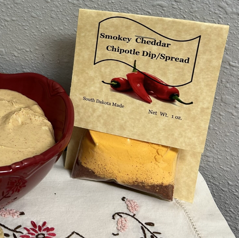 Smokey Cheddar Chipotle Dip/Spread package with a small bowl of dip on a floral tablecloth.