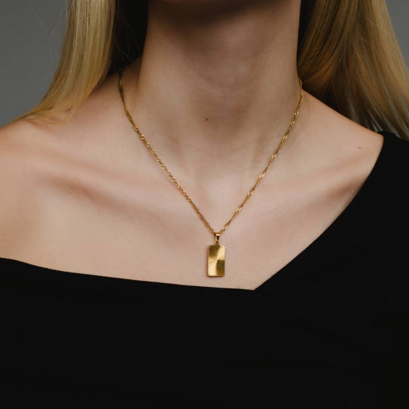 Gold necklace with a tag pendant worn by a person against a neutral background