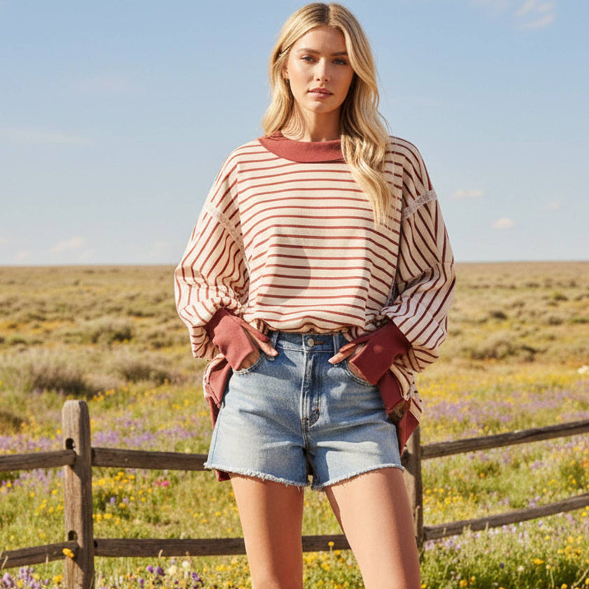 Woman standing in a field with wildflowers and a wooden fence, wearing a striped shirt and denim shorts.
