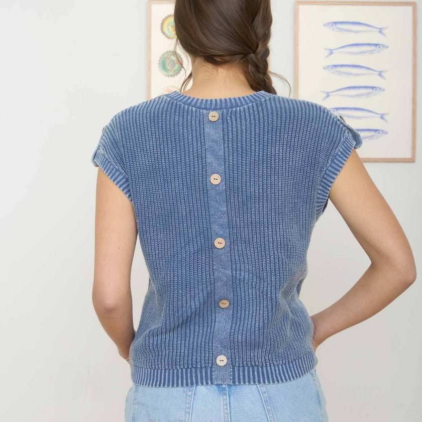 Woman wearing a blue ribbed knit sleeveless sweater with button details, standing in front of framed artwork on a white wall.