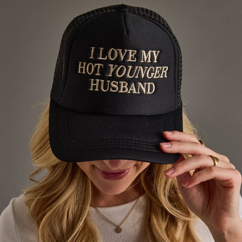 Person wearing a black cap with gold text, holding it over their face against a gray background