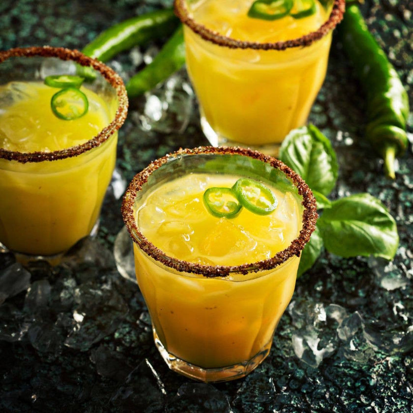 Three yellow cocktails with brown rims and jalapeño slices on a dark surface with ice.