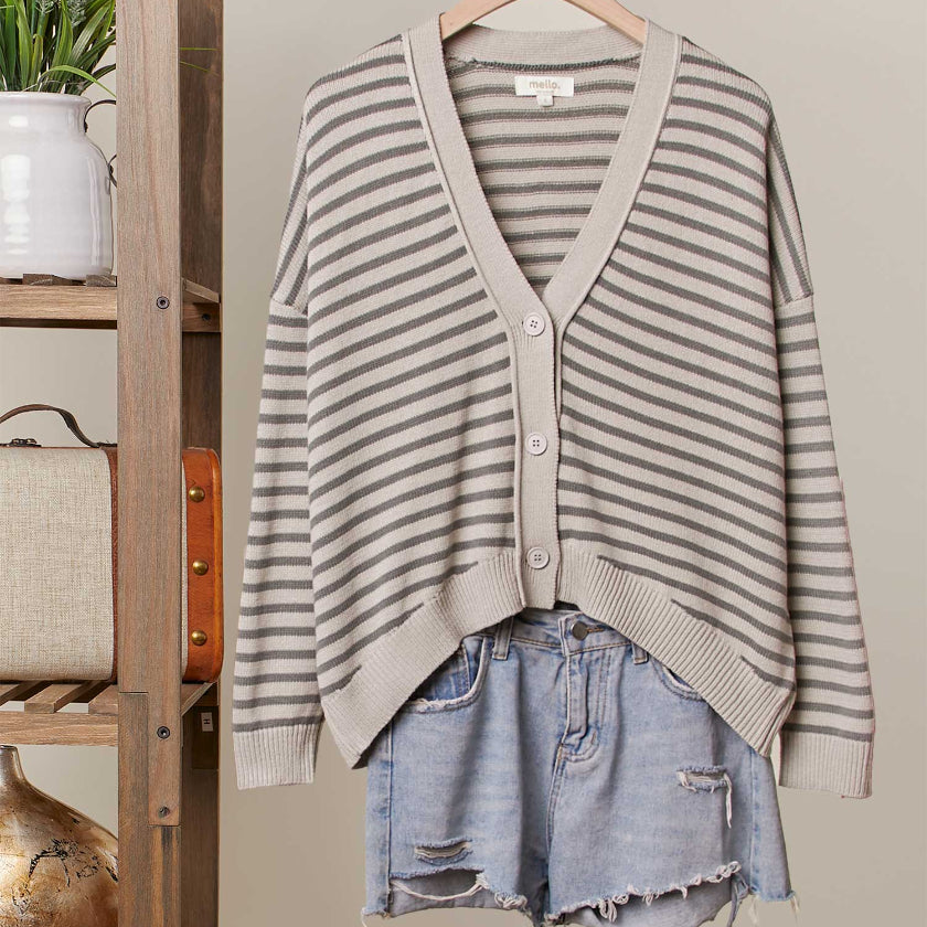 Striped cardigan and denim shorts on a hanger against a neutral wall with a wooden shelf and vintage suitcase.
