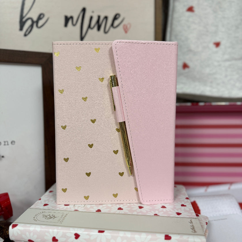 Pink notebook with gold heart patterns on a stack of books with heart designs.